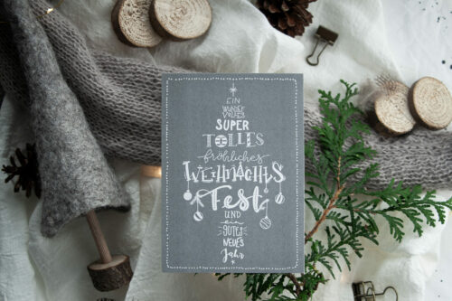 Weihnachtspostkarte "Lettering Weihnachtsbaum" im A6-Format mit handgezeichnetem Lettering-Weihnachtsbaum auf grauem Hintergrund, gedruckt auf 300g/m² Recyclingkarton. Die Karte zeigt weiße Schriftzüge, die einen Weihnachtsbaum formen, umgeben von kleinen weihnachtlichen Illustrationen wie Kugeln und Sternen. Dekoriert mit winterlichen Elementen wie Tannenzweigen, Kiefernzapfen und Holzscheiben.