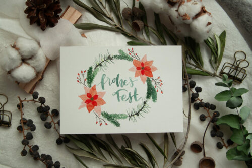 Weihnachtspostkarte im A6-Format mit handgemaltem Aquarell-Kranz aus Tannenzweigen, roten Weihnachtssternen und Beeren. In der Mitte steht der Schriftzug „Frohes Fest“. Die Karte ist auf hochwertigem Recyclingkarton gedruckt und ideal für nachhaltige Weihnachtsgrüße. Gestaltet von Frau Feierlich