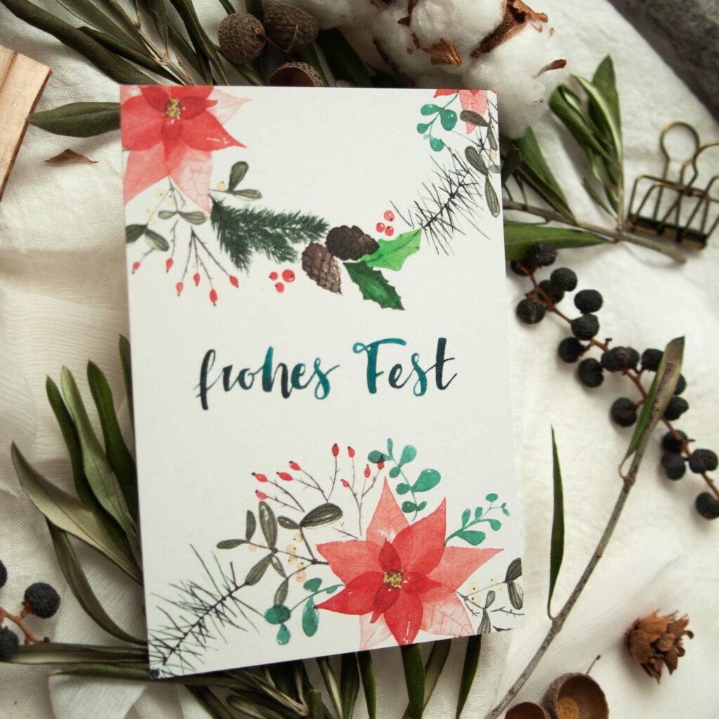 Weihnachtspostkarte mit handgemaltem Aquarell-Design, das Tannenzweige, Weihnachtssterne, Stechpalmen und Tannenzapfen zeigt, mit dem Schriftzug „Frohes Fest“. Gestaltet von Frau Feierlich