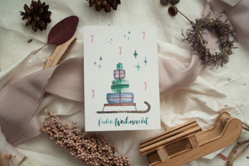 Weihnachtspostkarte mit handgezeichnetem Aquarell-Motiv eines Schlittens, beladen mit Geschenken und weihnachtlichen Verzierungen wie Sternen und Zuckerstangen, dazu der Schriftzug „frohe Weihnacht“ in festlichem Grün.Gestaltet von Frau Feierlich